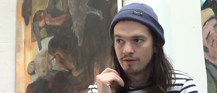 headshot photo of a young ethnically-ambiguous man with long hair, a blue beanie and facial hair in front of a painting in browns and beiges