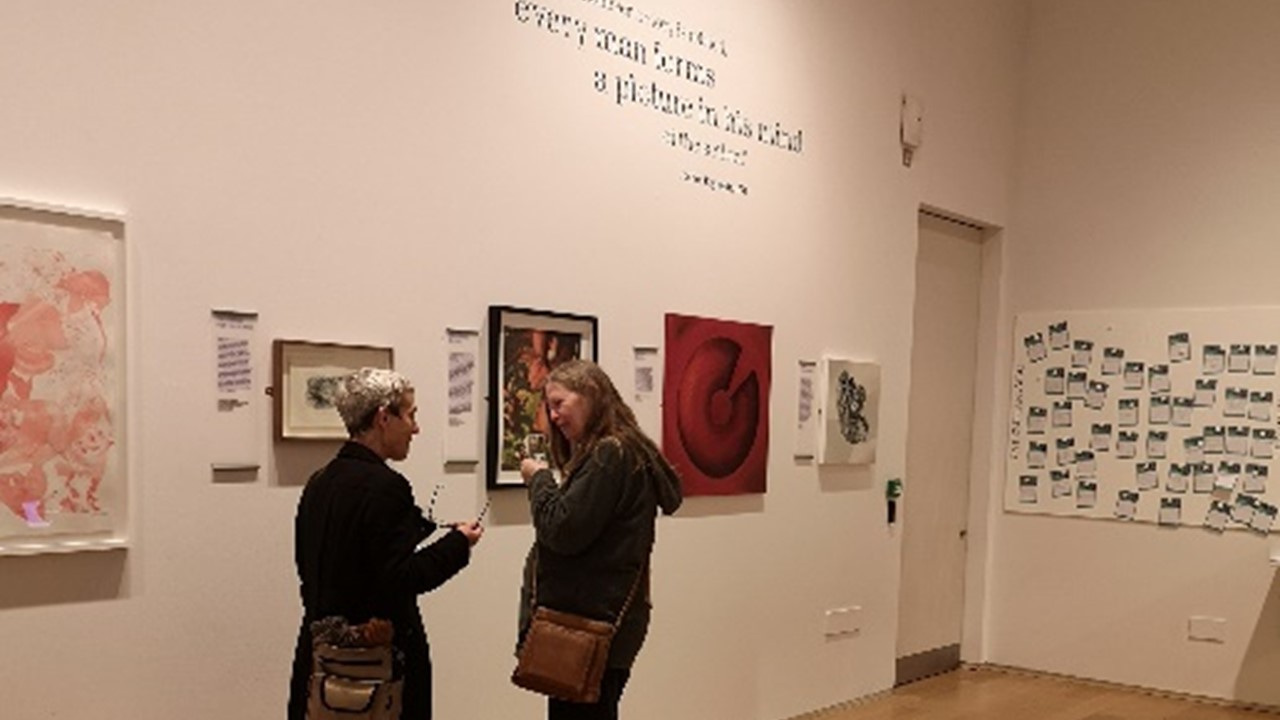photo of two white women deep in conversation, against a white wall of a gallery full of artwork that we can see in the online exhbition, as well as description writing in black on the wall. On the back wall in the background, comment cards are attached to a felt panel.