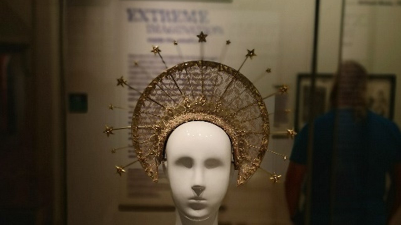 semi-dark photograph of one of the exhibits in the Extreme Imagination exhibition presented as a white mannequin bust with an elaborate golden crown that looks like a halo with golden spikes and stars offset from the edge of the halo. In the background, partly obscured, exhibition explanatory black text on a white wall