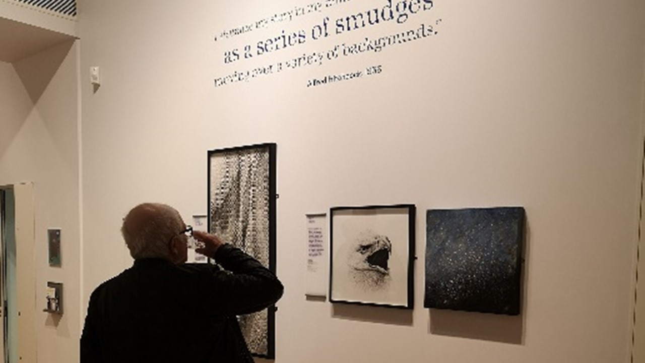 a photograph of the interior of the exhibition with a figure with the back towards the viewer, raising his hand as to shield his eyes or take a drink. In background two images that can be viewed in the online exhbition (a black and white textile and a blue sparkling non-figurative painting) as well as the charcoal drawing of an eagle hang framed on a wall.