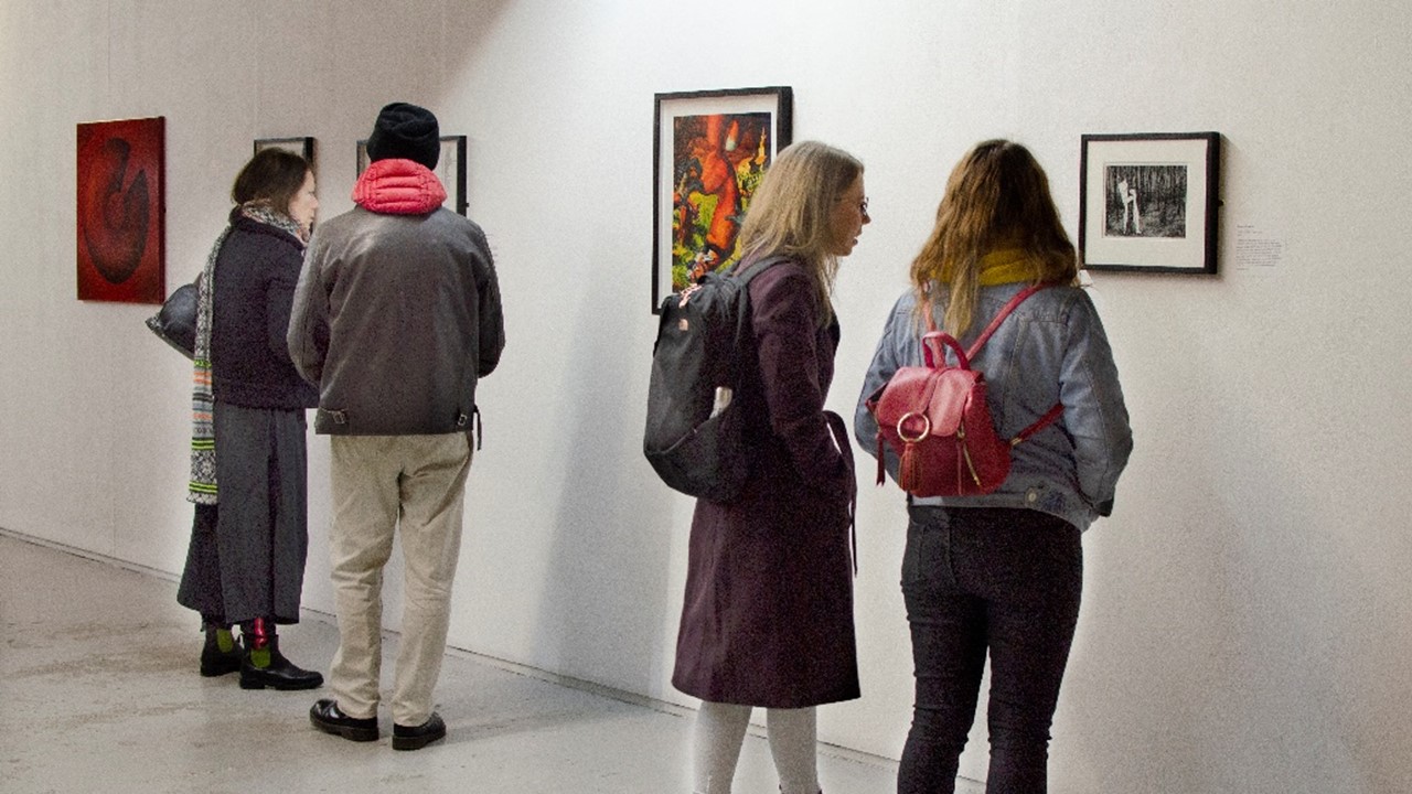 long panoramic view of the inside of the exhibition with four figures of different genders in front of white walls with artwork framed and hung up.