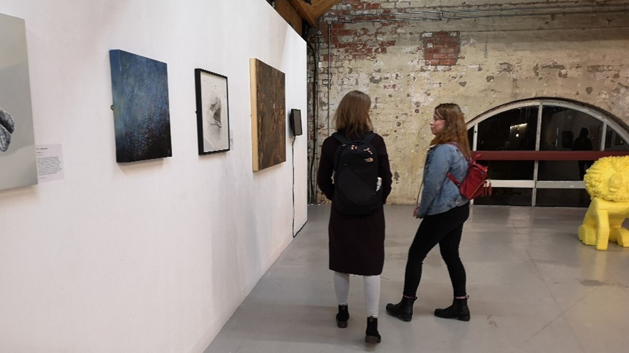 long panoramic view of the inside of an exhibitoon space with one long white wall with artwork framed and hung and a back wall made out of stone with a vaulted window framed by darker blocks of stone. Two people talk to each other, partially facing the artwork hung on the wall