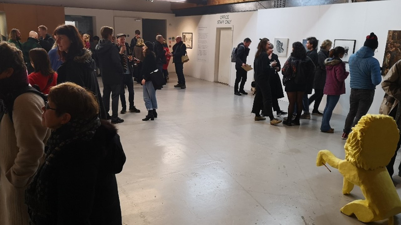 wide image of an unconventional gallery space with white as well as stone walls, with a lot of people in small groups looking at pictures on the wall and discussing. In the foreground we can see the yellow cast lion