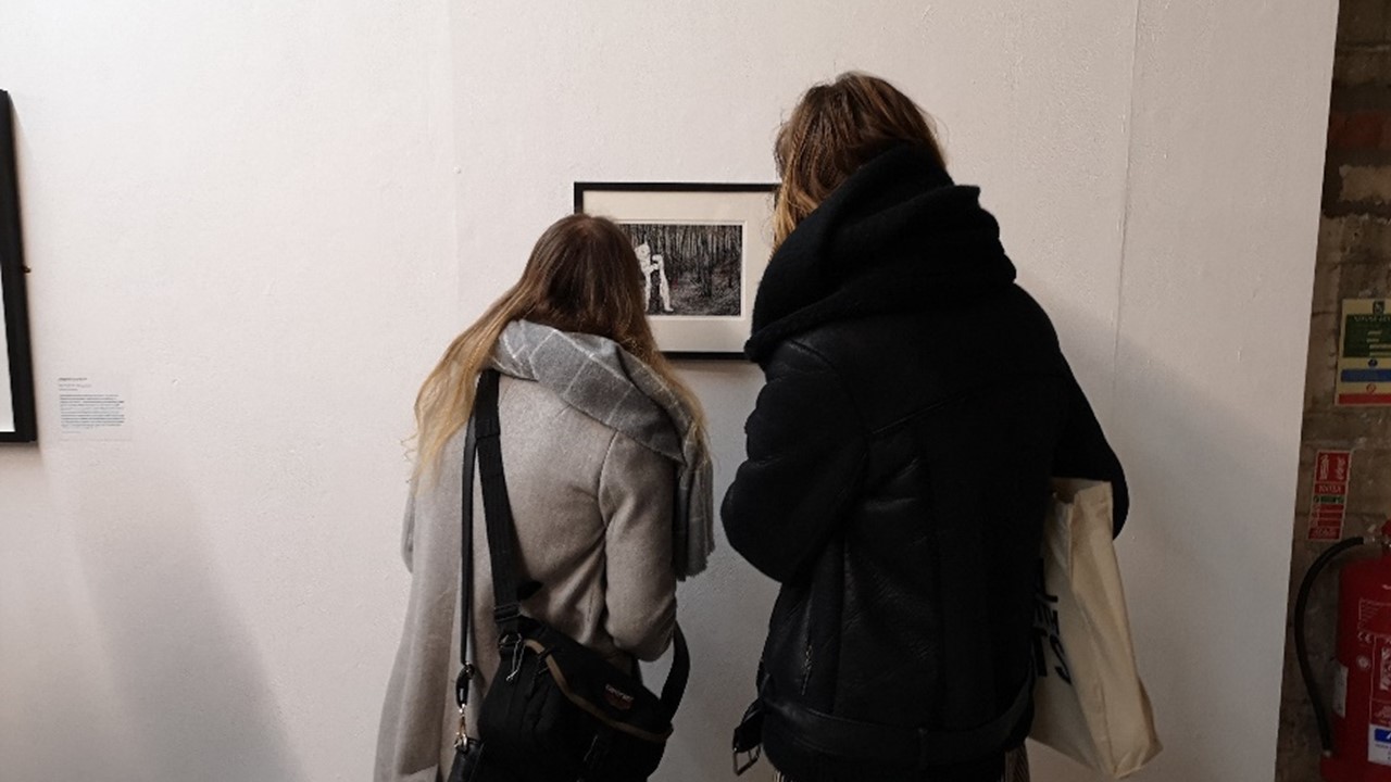 photo of two people in puffy jackets gathered close around an artwork framed and hung on a white wall. The artwork is a drawing print of a polar bear in the woods.